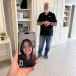 A hand holds a smartphone in the foreground showing a video call with a smiling woman, while a man stands blurred in the background of a bright, modern home interior with white Murphy Beds and tiled floors.