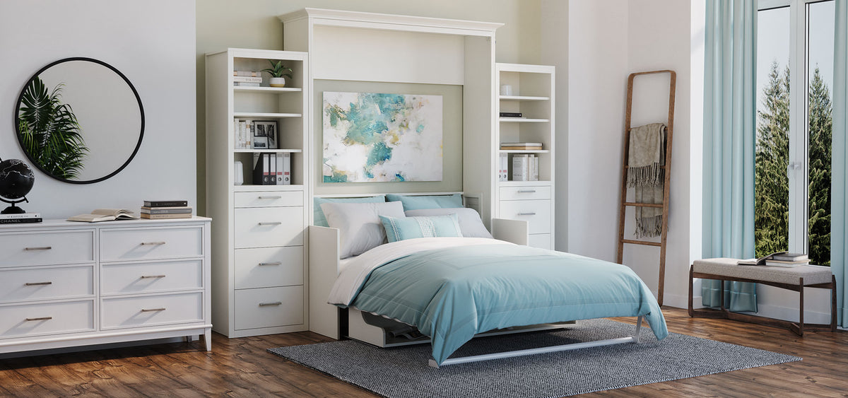 A white wall-mounted Murphy bed frame in a bright bedroom, shown with a light blue and white bedding set. The bed is flanked by tall white storage shelves, with a matching white dresser on the left and a window with sheer curtains on the right.