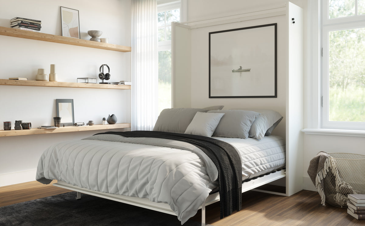 A modern white Murphy bed in a bright, minimalist bedroom. The bed is open to show a light gray quilted bedding set with a black throw blanket.